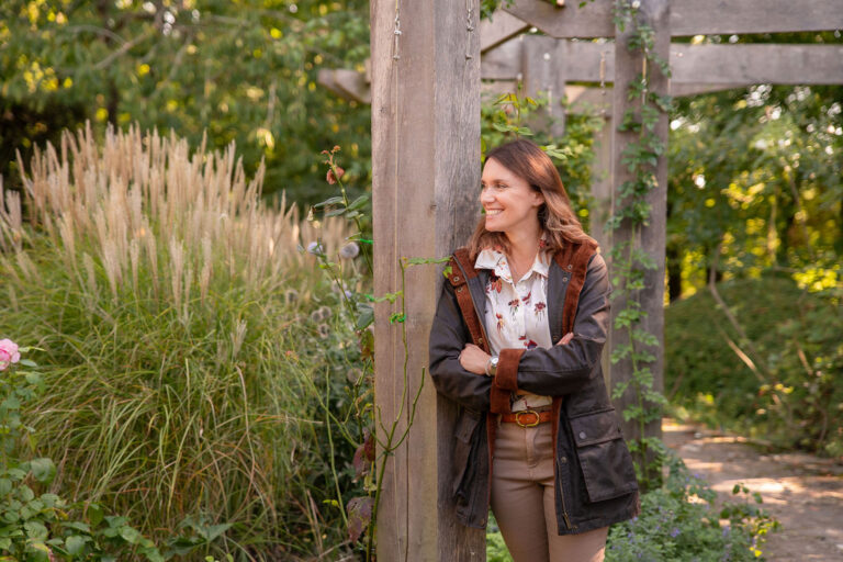 Garden Designer Victoria Rumens leaning against a wooden arbour surrounded by ornamental grasses.