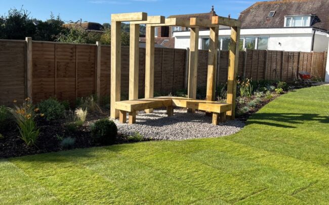A fire pit area with gravel and wooden bench and structure for climbing plants in a garden in Findon Valley