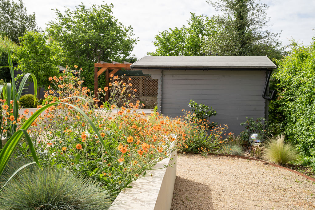 Orange geum and grasses spilling over a rendered wall beside a gravel path